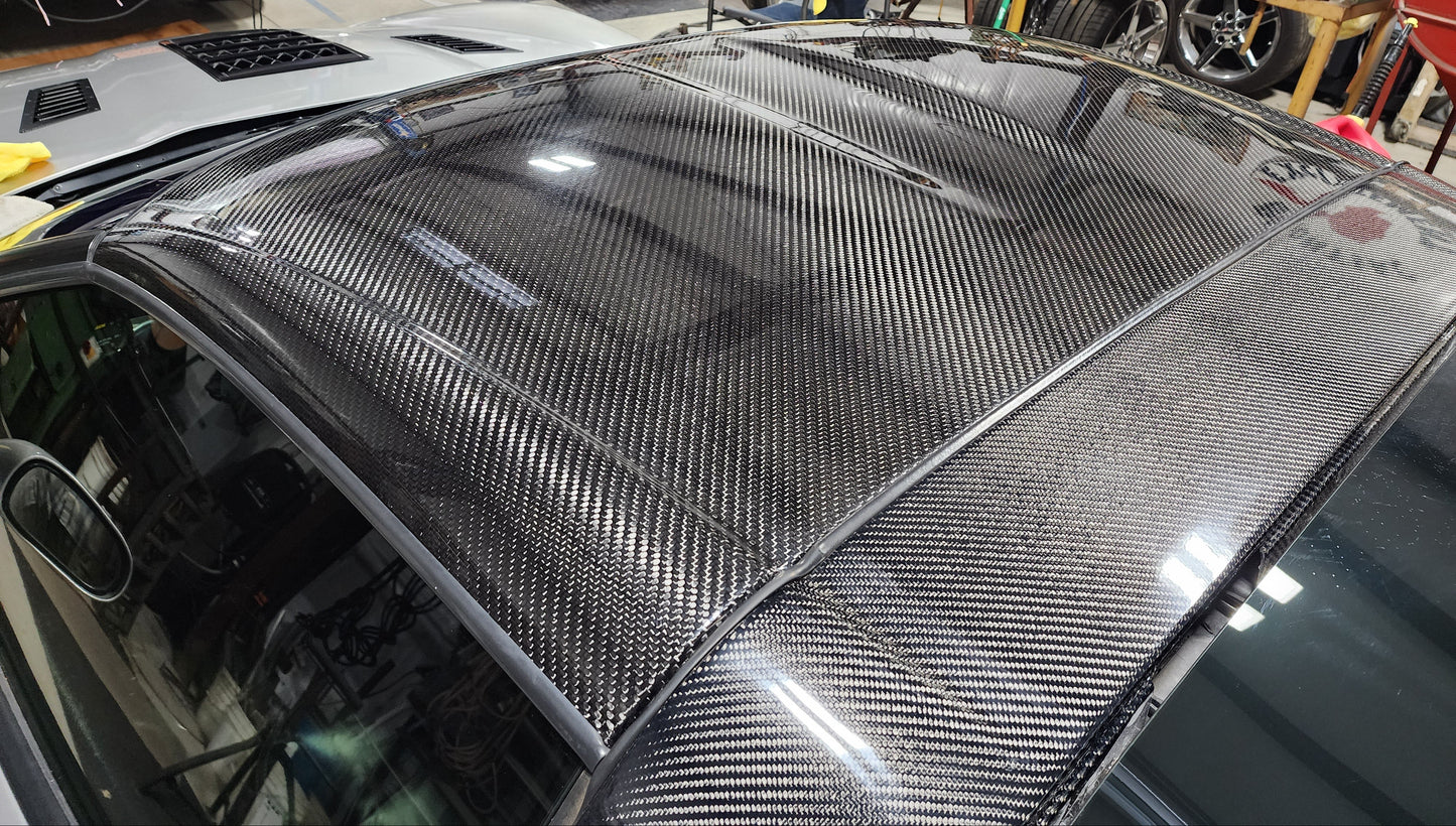 A carbon fiber car roof panel with a glossy woven carbon fiber pattern, installed on a vehicle in a workshop environment.