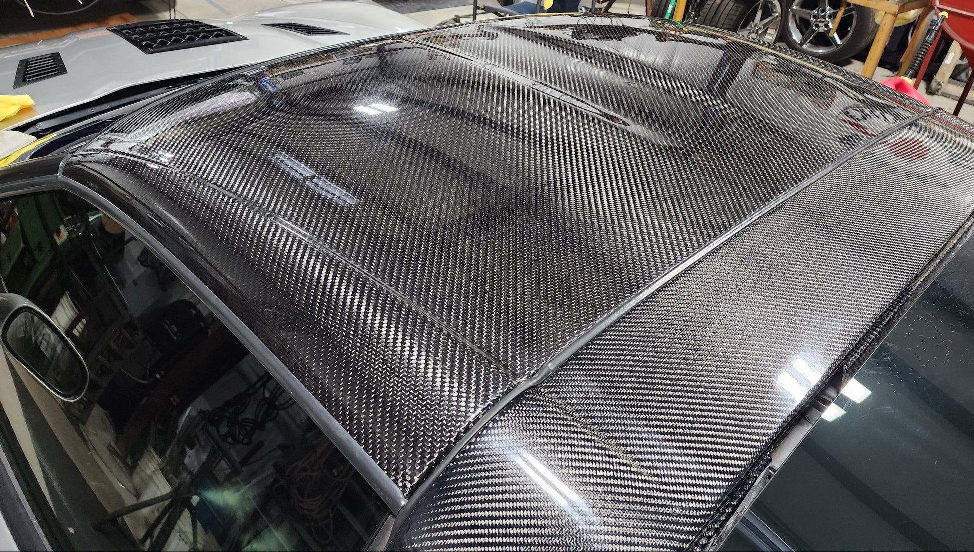 A carbon fiber car roof panel with a glossy woven carbon fiber pattern, installed on a vehicle in a workshop environment.