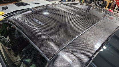 A carbon fiber car roof panel with a glossy woven carbon fiber pattern, installed on a vehicle in a workshop environment.