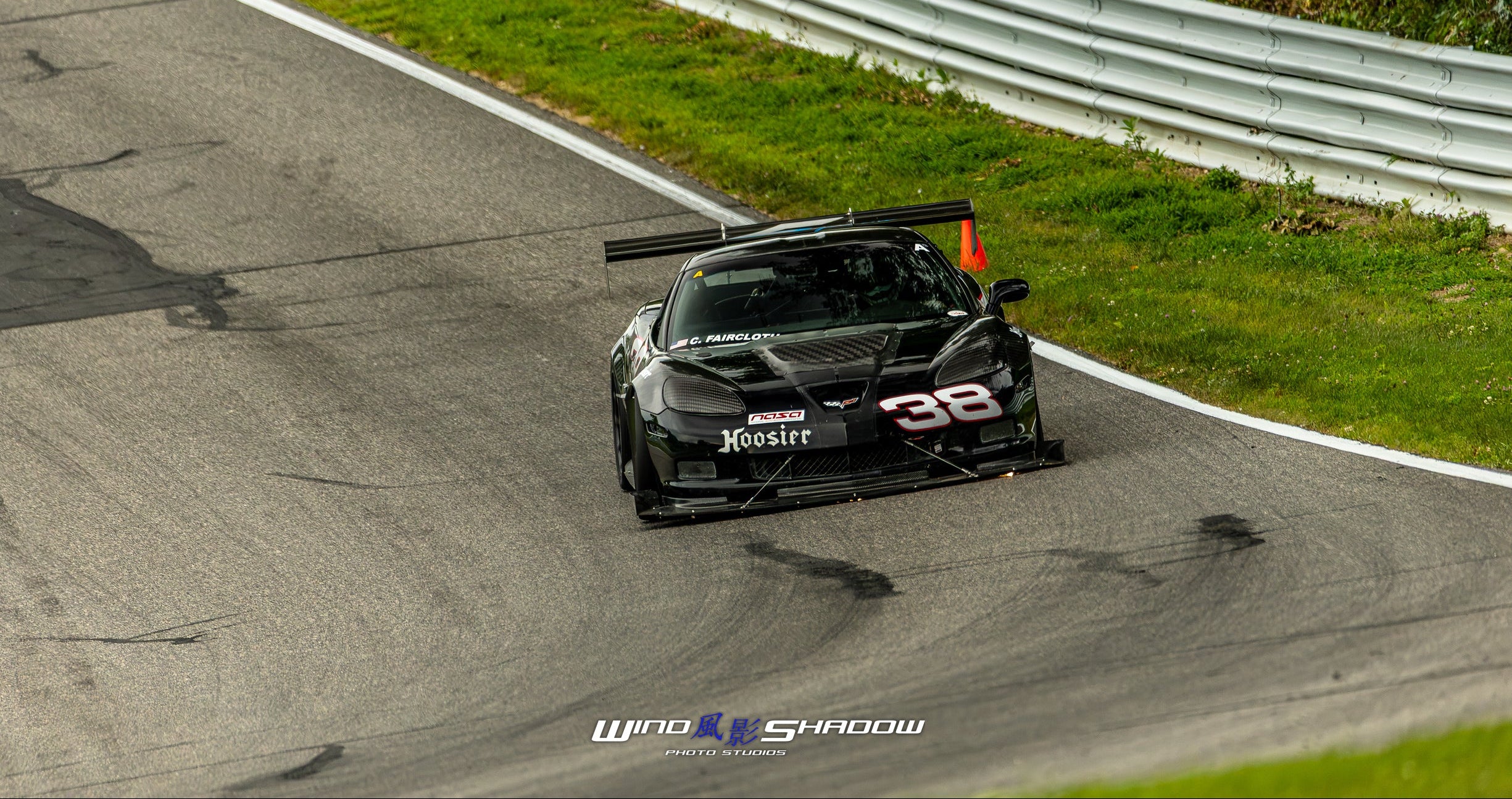 A black race car with the number 38 on the front, photographed from above on a racetrack with grass, guardrail, and wet asphalt visible. The photo includes a Wind X Shadow watermark at the bottom.