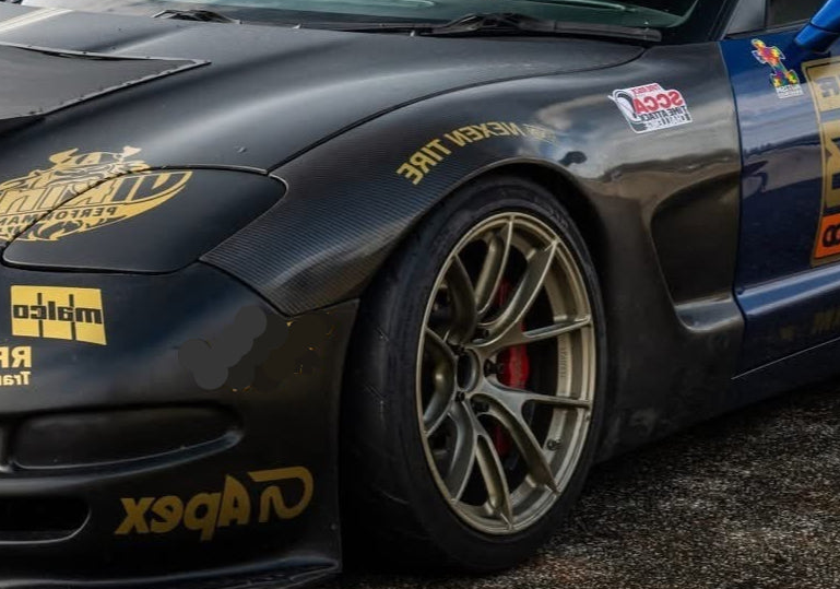 Close-up of a dark carbon-fiber race car body with gold sponsor decals, including Apex, Viking performance, and Nexen Tire, and a bronze multi-spoke wheel.