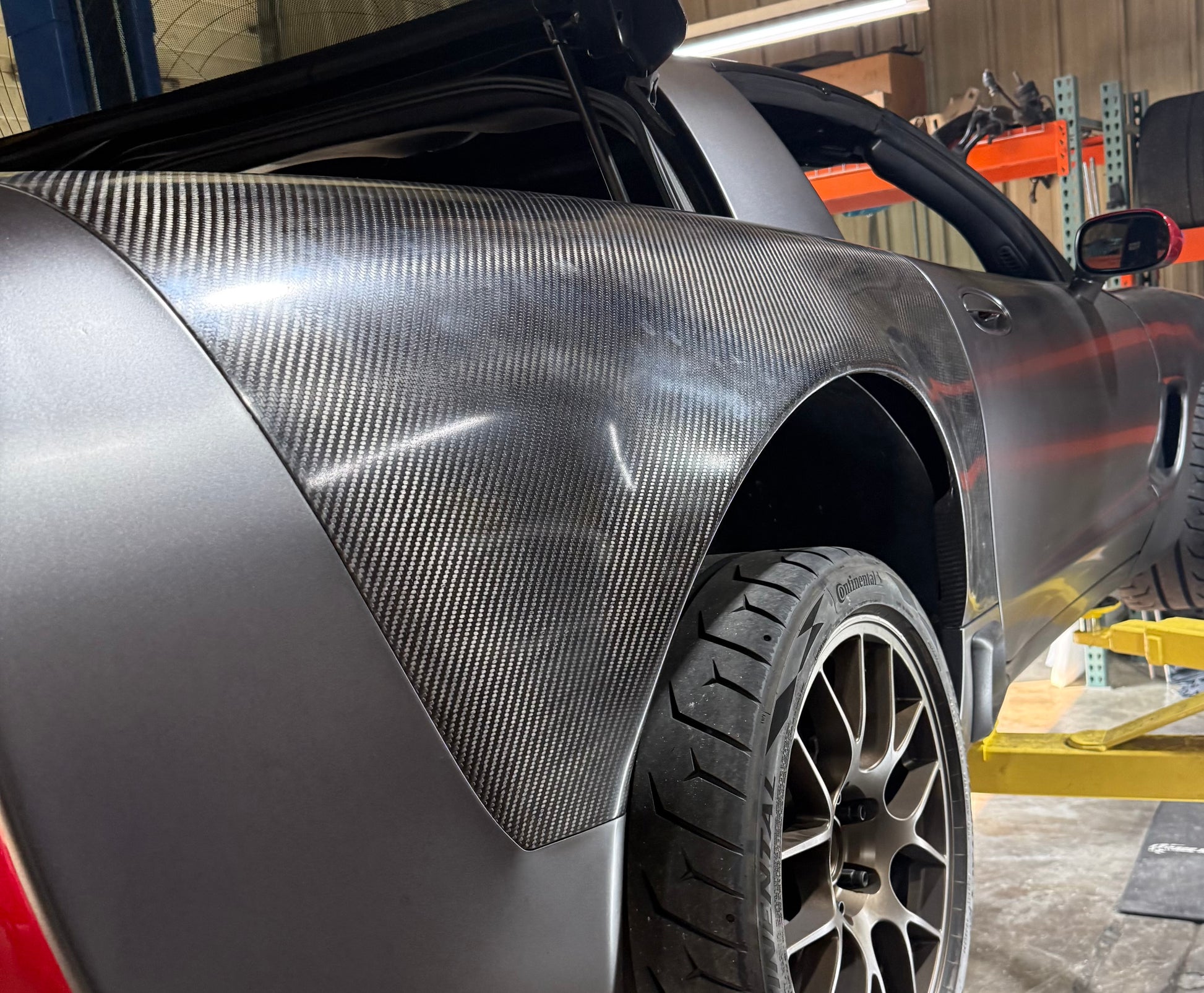 A close-up of a dark gray Corvette with a carbon fiber rear fender panel installed, photographed in a workshop setting.