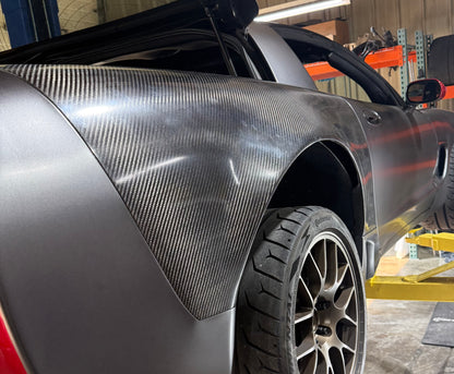 A close-up of a dark gray Corvette with a carbon fiber rear fender panel installed, photographed in a workshop setting.
