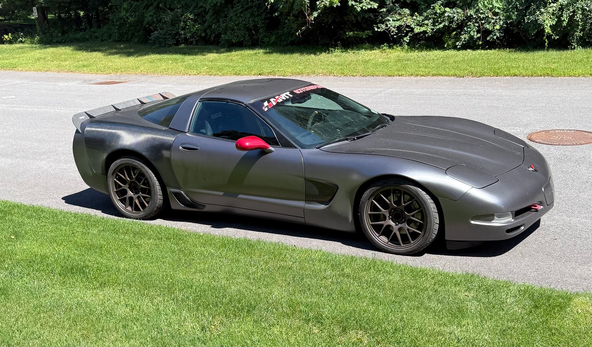 A dark gray Corvette sports car parked on an asphalt driveway with green grass and trees in the background.