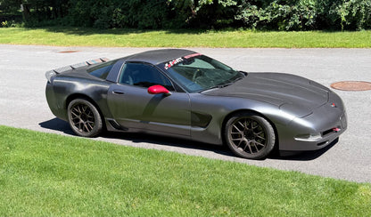 A dark gray Corvette sports car parked on an asphalt driveway with green grass and trees in the background.