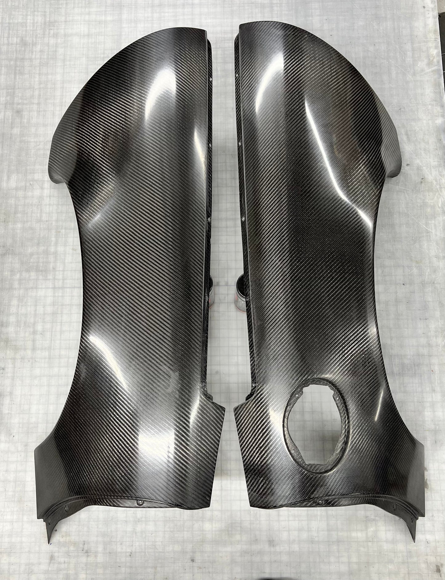 A pair of glossy black carbon fiber body panels with a visible woven carbon fiber pattern, displayed on a light gray textured surface.