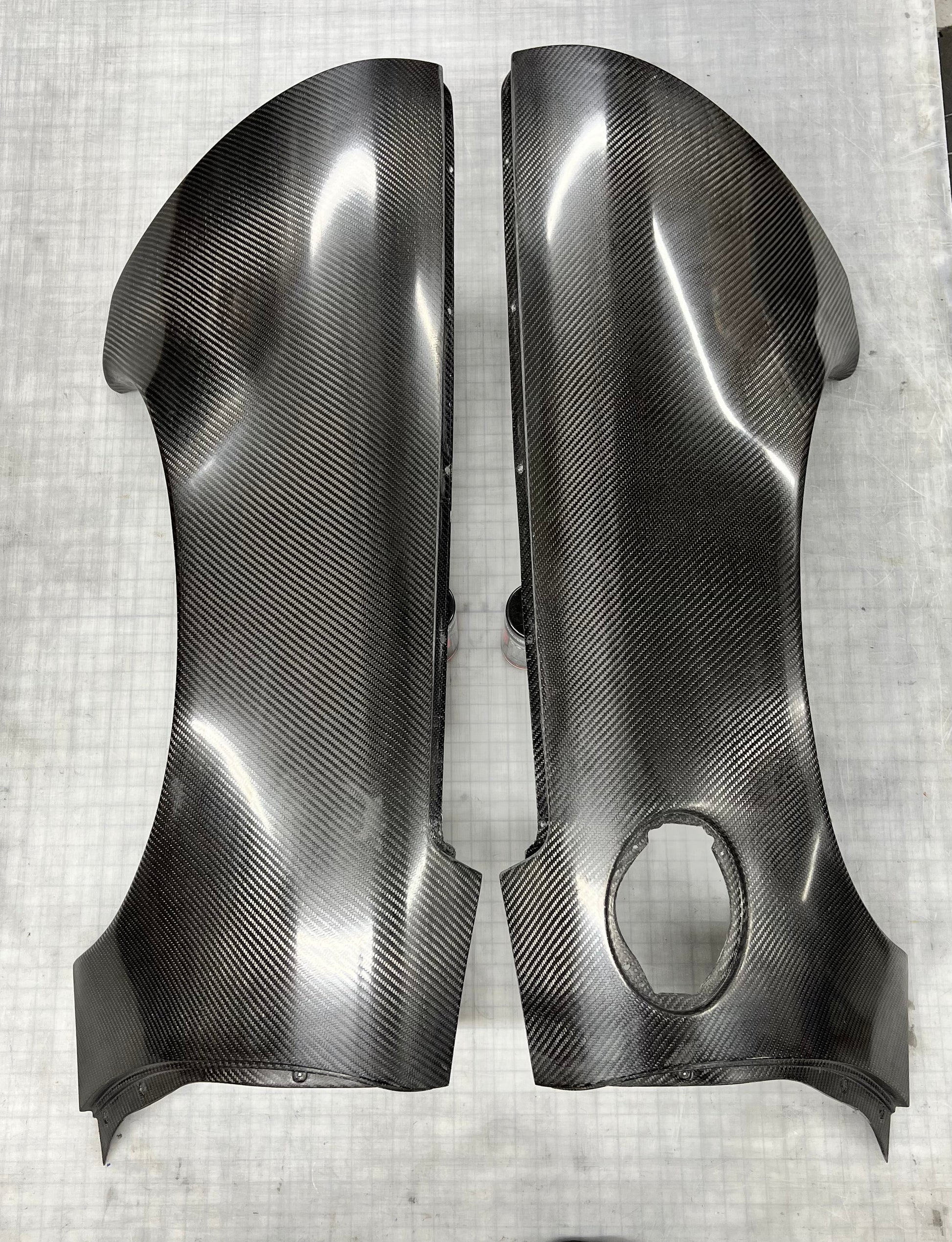 A pair of glossy black carbon fiber body panels with a visible woven carbon fiber pattern, displayed on a light gray textured surface.