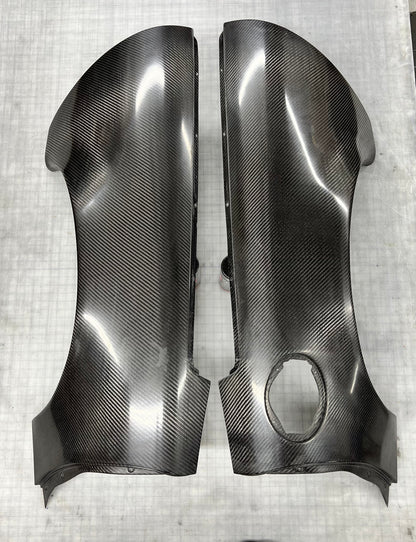 A pair of glossy black carbon fiber body panels with a visible woven carbon fiber pattern, displayed on a light gray textured surface.
