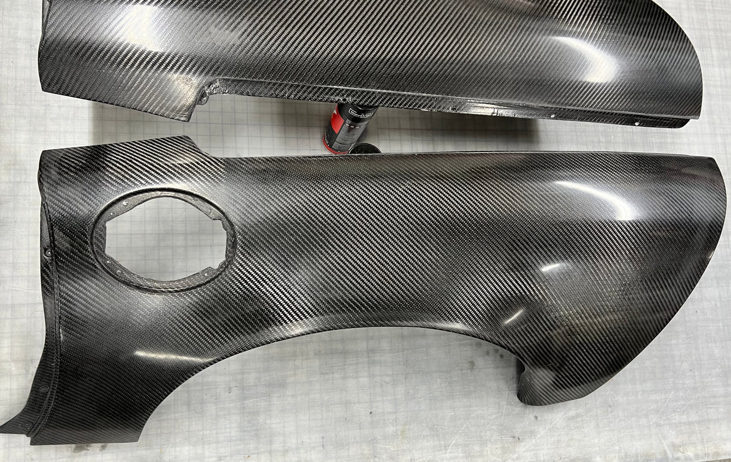 Two glossy carbon fiber body panels with a visible woven carbon weave pattern, photographed on a light gray textured surface.