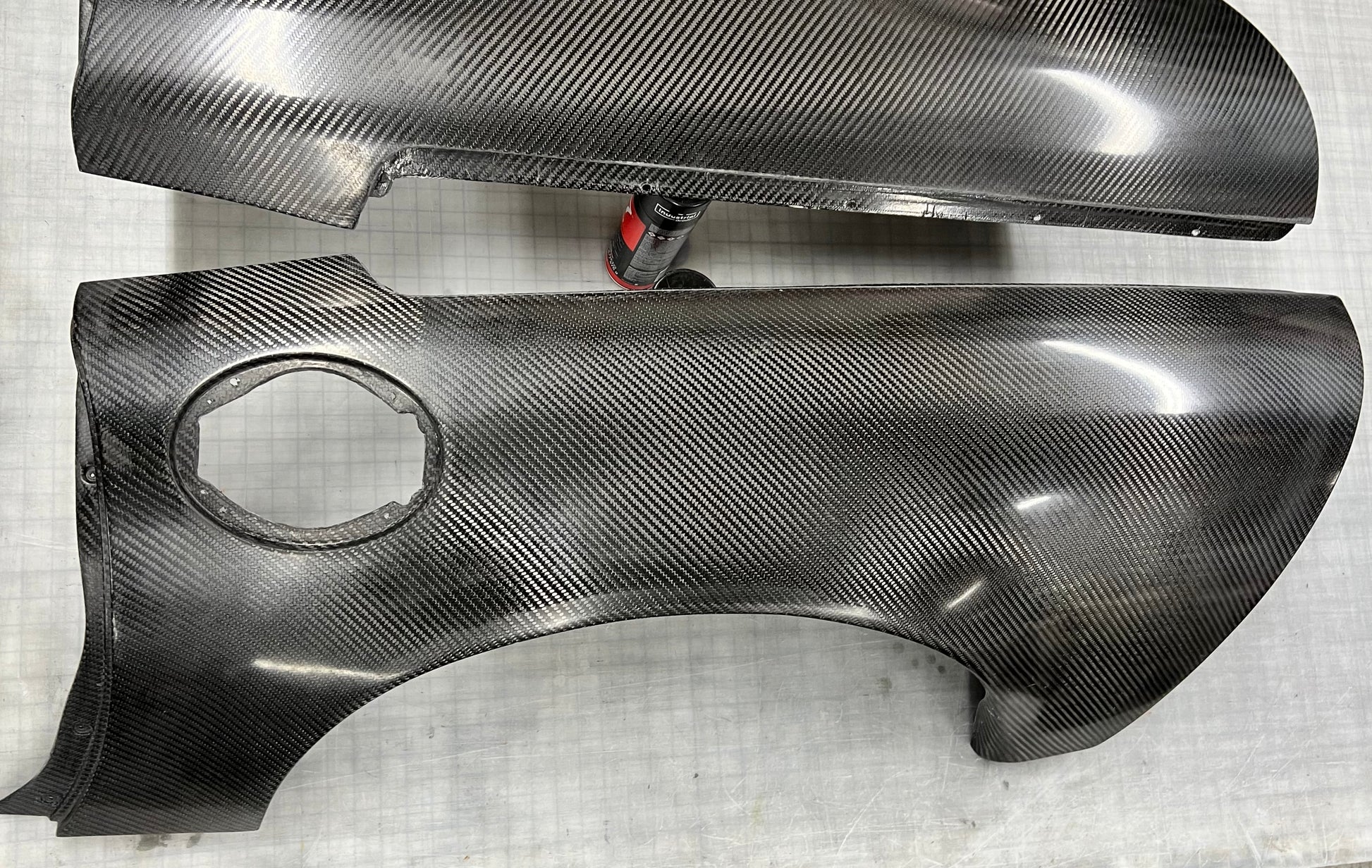 Two glossy carbon fiber body panels with a visible woven carbon weave pattern, photographed on a light gray textured surface.