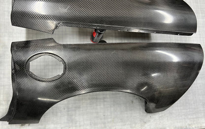 Two glossy carbon fiber body panels with a visible woven carbon weave pattern, photographed on a light gray textured surface.