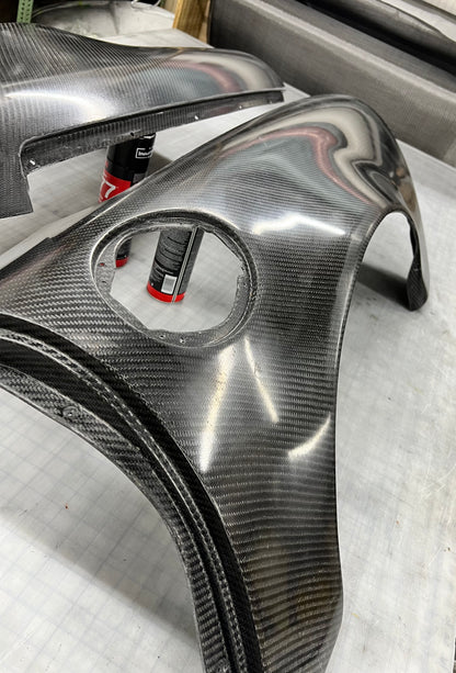 A glossy carbon fiber automotive body panel with a visible woven carbon pattern, photographed on a light workbench with other matching parts in the background.