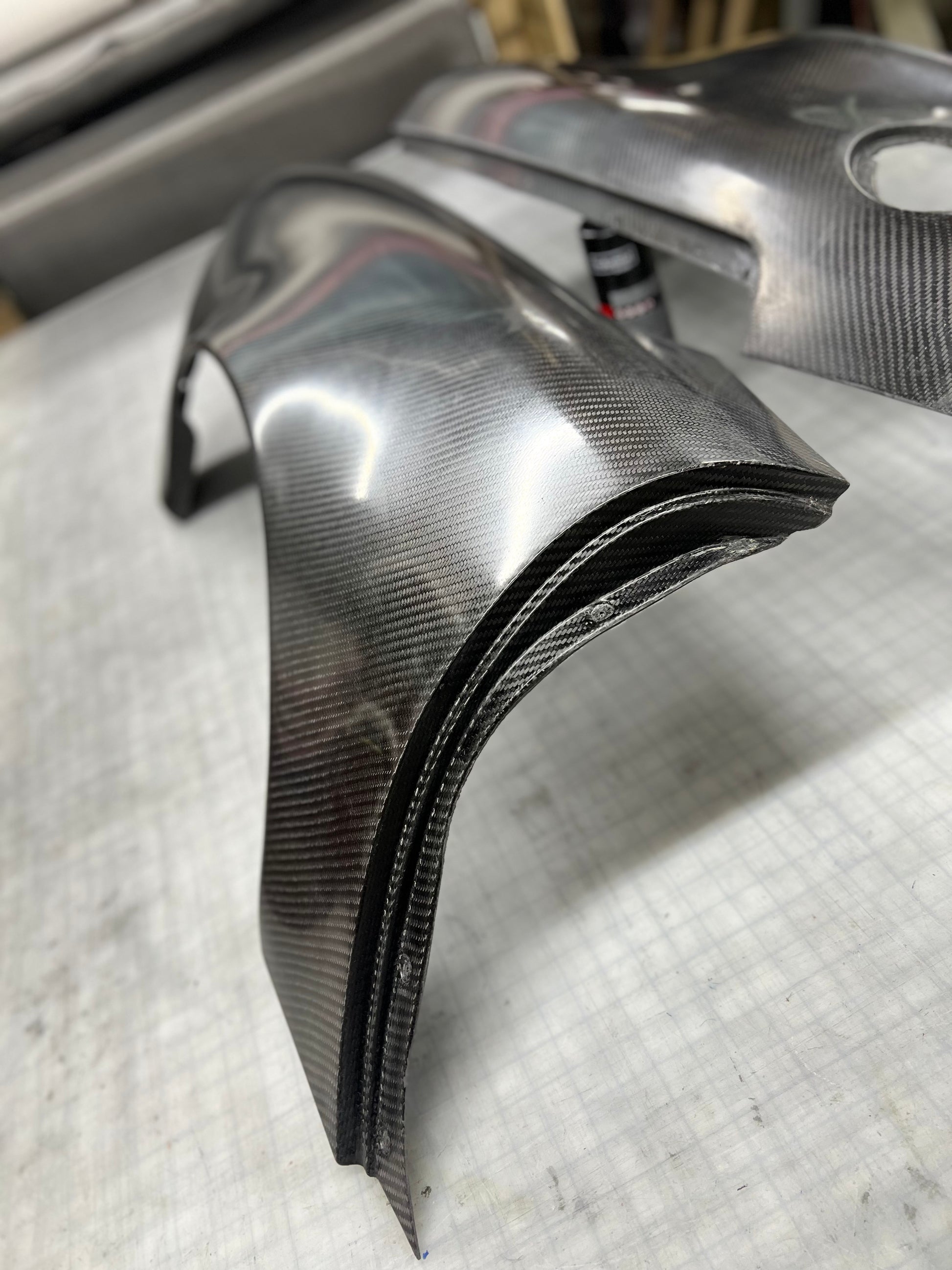 A pair of custom carbon fiber body panels, likely fenders or trim pieces, displayed on a gray workbench. The panels feature a glossy, high-contrast woven carbon fiber pattern with black edges.
