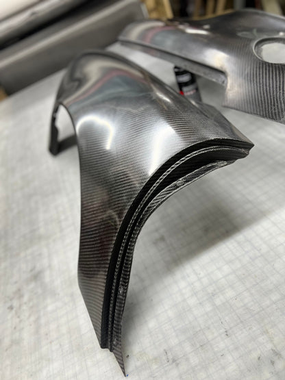 A pair of custom carbon fiber body panels, likely fenders or trim pieces, displayed on a gray workbench. The panels feature a glossy, high-contrast woven carbon fiber pattern with black edges.