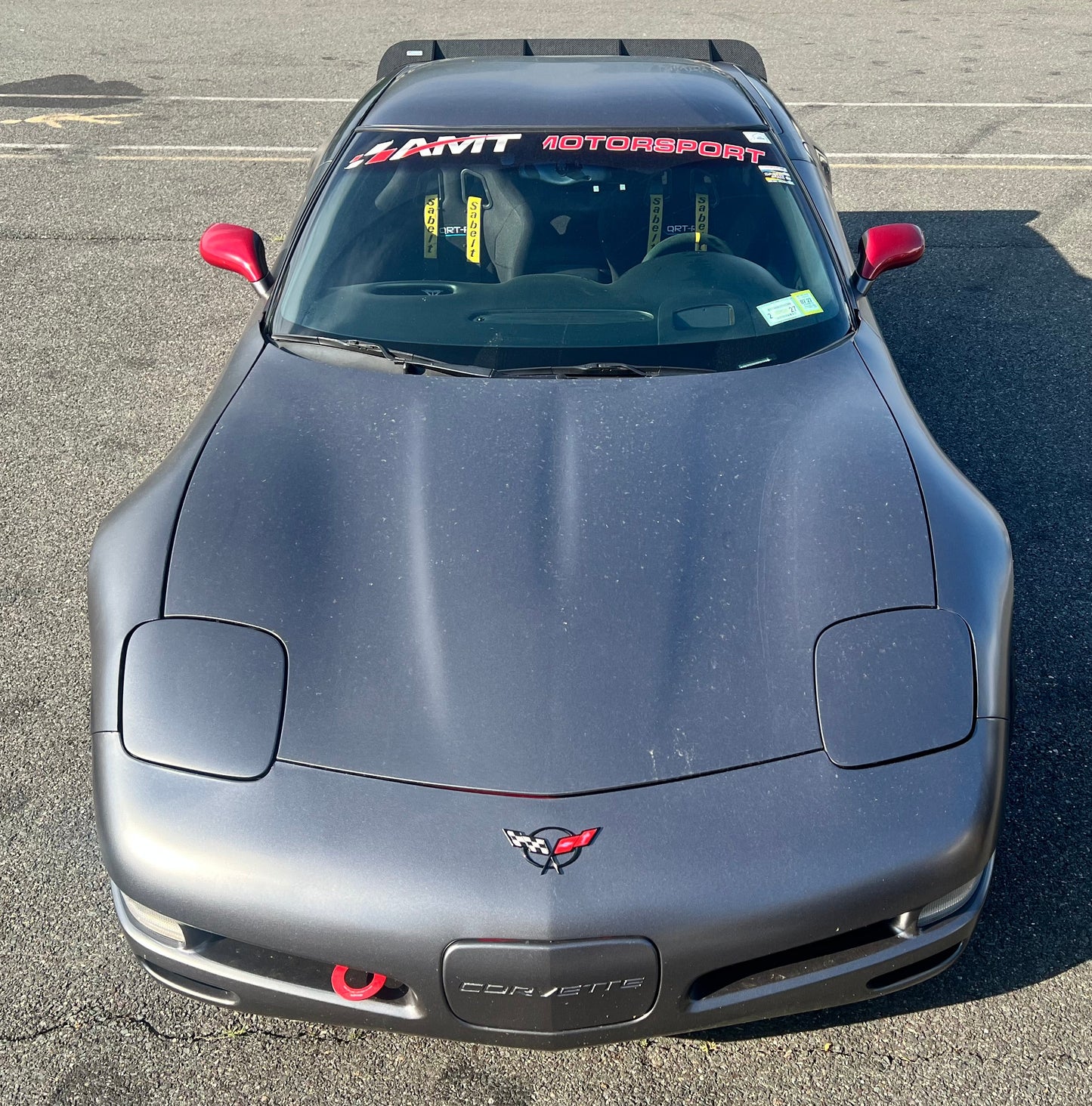 A dark gray Corvette C5 coupe race car with red side mirrors and a 'AMT Motorsport' windshield decal, photographed from the front in an outdoor paved lot.